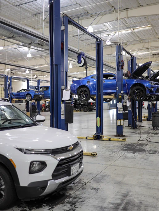 A car service area with several Chevrolet vehicles, including blue cars on lifts and a white car in the foreground. The garage has high ceilings, metal beams, and bright lighting.