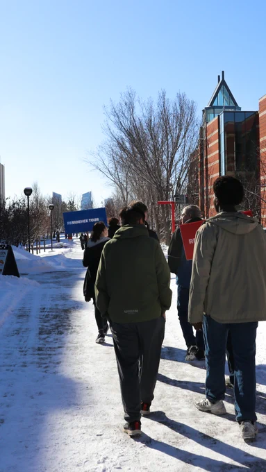 A group of people walk along a snowy path towards the SAIT campus, surrounded by bare trees and modern buildings.