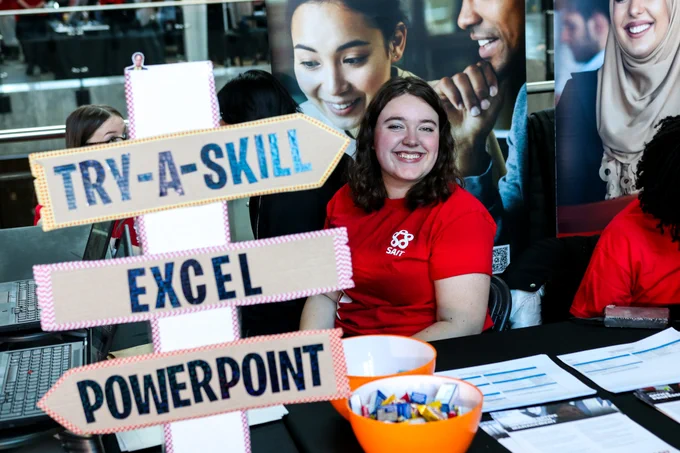 A person wearing a red shirt is sitting at a table with brochures and a bowl of candy. There is a sign in the foreground with arrows labeled "Try-A-Skill," "Excel," and "PowerPoint." Banners with smiling faces are visible in the background.