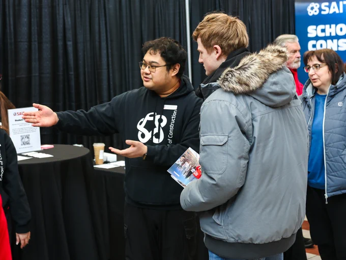 Two people are engaged in conversation at a booth during a conference. One person is gesturing with an open hand, and the other holds a brochure. The booth displays the SAIT logo and the words "School Cons". There are some items on a round table, including QR codes. Other attendees are visible in the background.