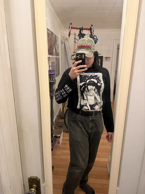 Me taking a mirror selfie with my phone kinda obscuring my face. I’m wearing moss green jeans, with a long sleeve black shirt tucked in. The shirt has a graphic of the Pigeon Pit album cover for Treehouse, a drawing of someone eating a peach, and the sleeves have lyrics (visible is “forget all the”). I’m wearing a hat the same color as my jeans with a red patch sewn on that says “puppygirl.”