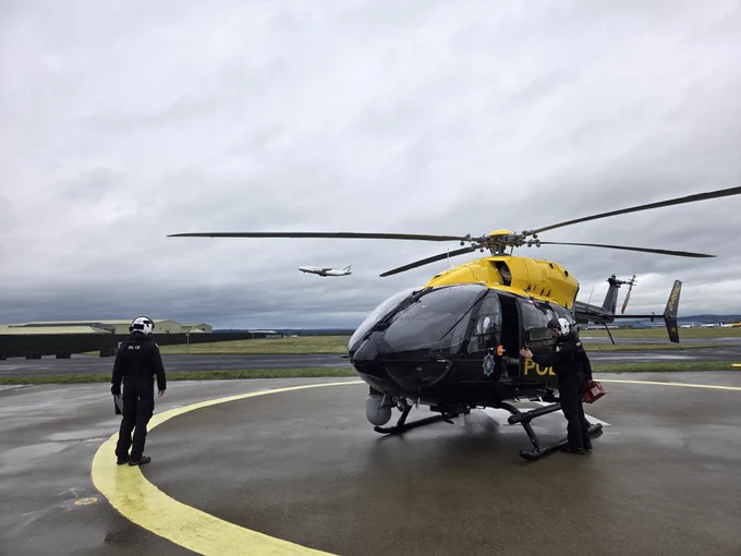 The Police helicopter crew are walking towards the police helicopter while an Antonov aircraft takes off in the background