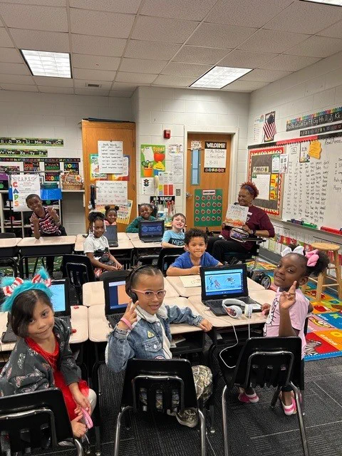Elementary students sitting at desks with laptops in a colorful classroom, engaging with a teacher holding a book.