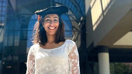 A woman in a white lace dress and graduation cap stands smiling in front of a modern glass building.