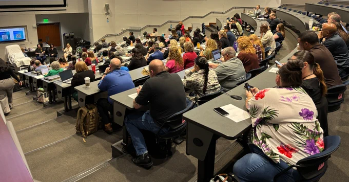 A large classroom filled with people seated at rows of desks facing the front, where multiple screens display Windows logos. A lecturer is speaking to the audience.