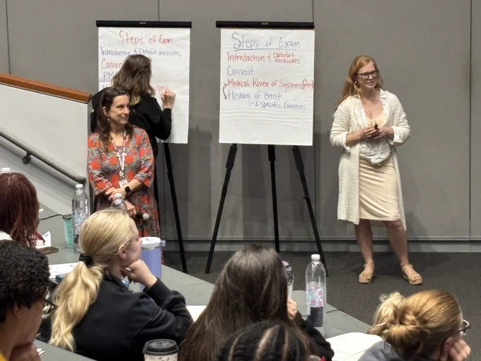 A classroom setting with two people standing next to flip charts at the front. The flip charts display step-by-step outlines: "Steps of Exam" and "Steps of Exam: Introduction, Consent & Measures, Medical Record & Systems (Prof), History of Present, Specific Questions." Several students are seated, facing the speakers, taking notes.