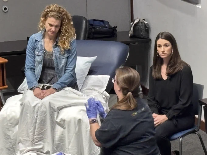 A person sits on a medical examination table while two others, one in a black top and another with gloved hands, are nearby. The atmosphere suggests a medical consultation setting, with a focus on the person in blue denim on the table.