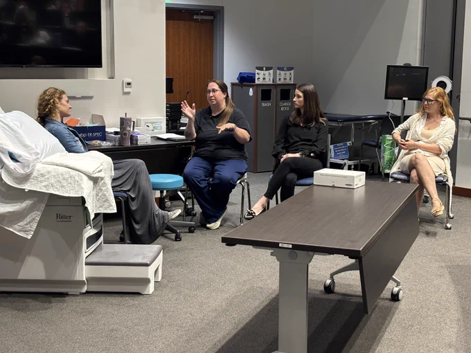 A group of people are sitting in a room set up with medical equipment and a large monitor. The central figure is speaking while others listen attentively. One person is seated on a hospital bed. The atmosphere is classroom learning environment.