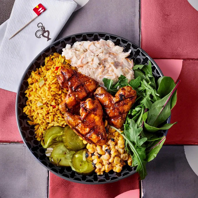 A bowl with grilled chicken, yellow rice, salad greens, pickles, creamy coleslaw, and corn on a bed of black and pink tiled surface. A napkin features the Nando's logo.