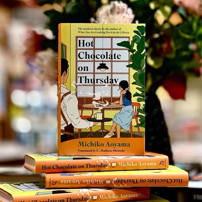 The image shows a stack of books titled "Hot Chocolate on Thursday" by Michiko Aoyama, translated by E. Madison Shimoda. The cover features an illustration of two people sitting at a table with hot drinks. Flowers are slightly blurred in the background.