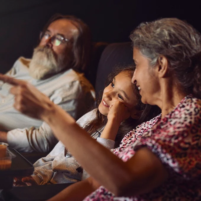 A group of three people sit closely in a dimly lit theater, attentively watching a screen. The middle person, with long hair, looks delighted, resting their chin on their hand. The person on the right gestures towards the screen, while the person on the left, with long hair and glasses, watches intently. They sit side by side in comfortable seats.