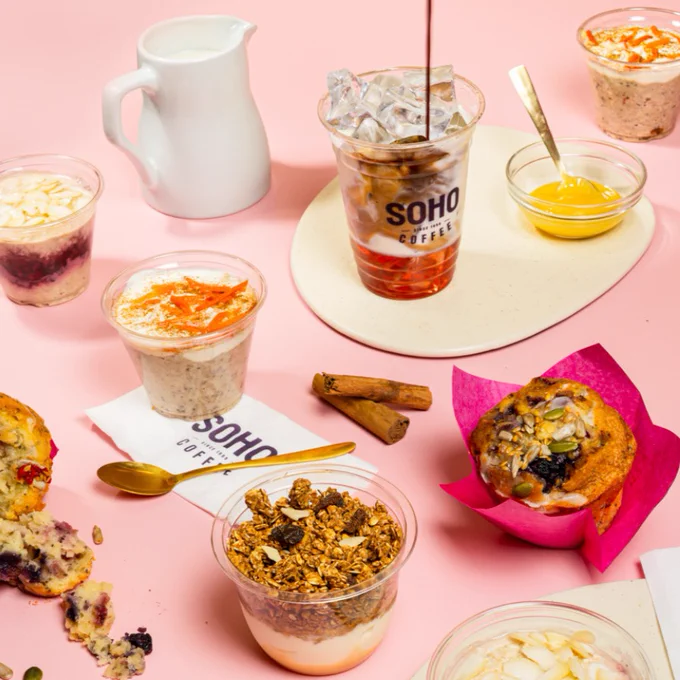 Various breakfast items are arranged on a pink surface, including yogurt parfaits, muffins, and a container of iced coffee with the SOHO Coffee Co. logo. A small jug of milk and cinnamon sticks are also present, with a mix of toppings and utensils scattered around.