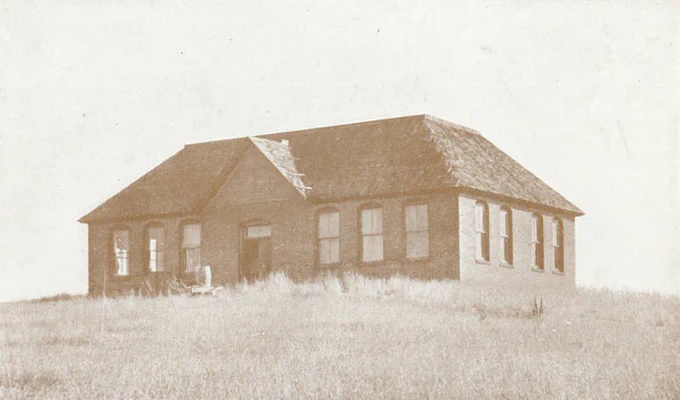 An old, single‑story brick building sits alone in an open grassy field. The structure has a steep, shingled roof with visible wear and damage, including a section where shingles are missing. Several tall, narrow windows line the sides, many appearing dark or empty. The overall scene looks aged and weathered, giving the impression of an abandoned rural schoolhouse or community building.