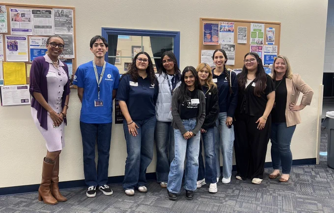 A group of nine diverse students and staff pose in a brightly lit office or classroom. Several wear blue AmeriCorps shirts. They stand in front of a blue-framed window and two bulletin boards covered in academic posters, including one for FAFSA. The group is dressed in casual business and student attire, ranging from jeans and t-shirts to a lavender dress with tall boots.