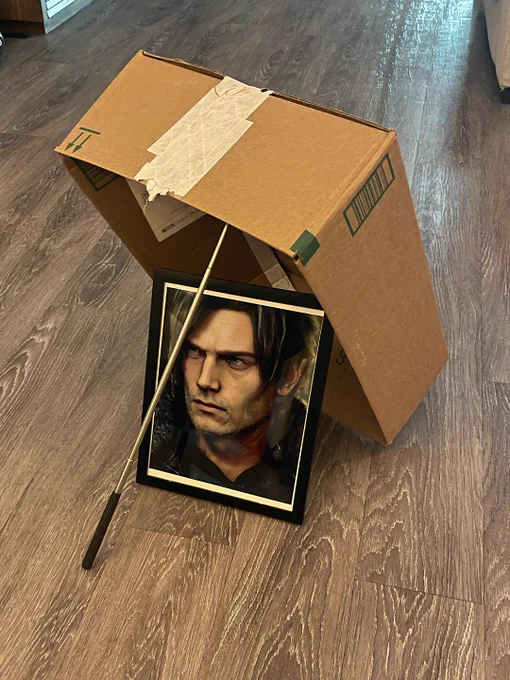 A cardboard box propped up with a stick like a classic trap sits on a wooden floor. Under the box is a framed photo of Leon from Resident Evil Requiem.