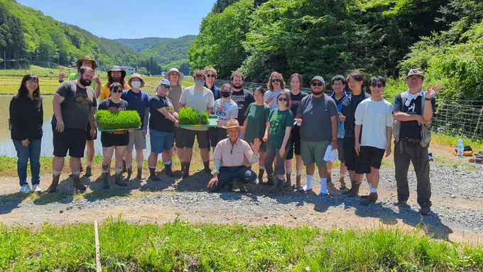 A diverse group of people stands on a gravel path in a scenic, rural area with a lush green landscape and mountains in the background. They are gathered around a person kneeling in front, holding trays of green plants. Everyone seems to be enjoying the bright, sunny day.