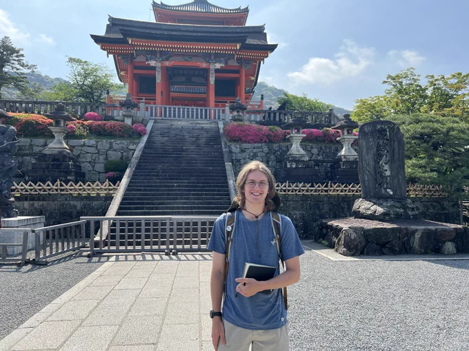 A person stands smiling in front of the Kiyomizu-dera, a historic temple in Kyoto, Japan. They are holding a tablet and wearing a backpack. The temple's red and white architecture is prominent, surrounded by lush greenery and bright flowers.