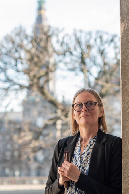 Diana står lænet op af en søjle ved ridebanen foran Christiansborg. I baggrunden kan Christiansborg slot anes. Diana har sin blindestok i hånden.