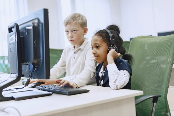 Two children in a computer lab using a desktop computer and headphones.