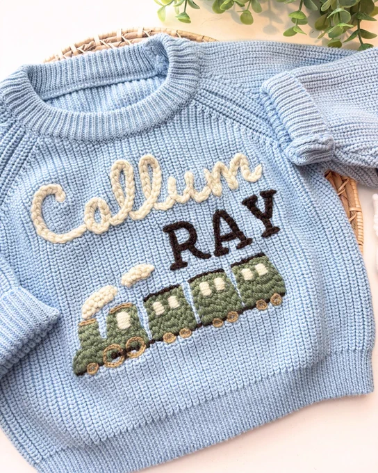 Light blue knitted sweater with a green train and the name Callum Ray embroidered on the front.