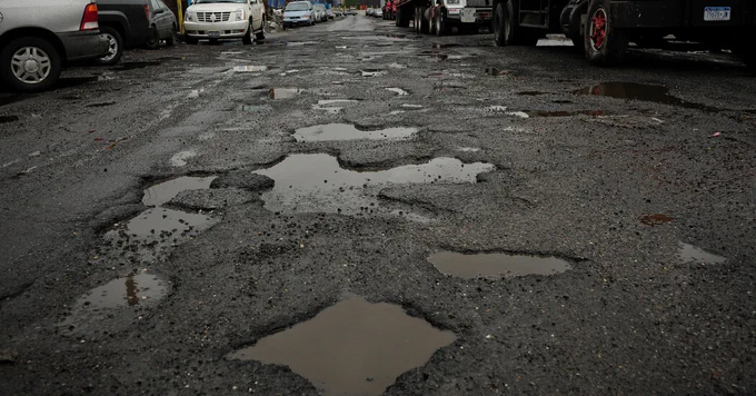 A road full of large potholes filled with water, surrounded by parked cars and trucks.