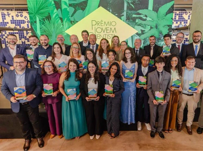 Fotografia retrata os vencedores do Prêmio Jovem Cientista 2025 de pé segurando seus troféus. Eles posam sorridentes em frente a um painel com a marca do prêmio. Sobre o fundo, formado por grafismos de árvores em tons de verde, há um losango amarelo-claro com o texto “Prêmio Jovem Cientista”.