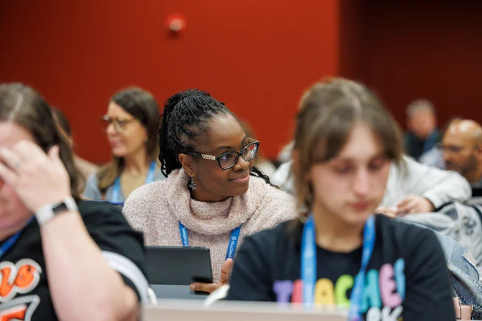A photo of a woman sitting down and looking to her left. She is in a room full of people sitting at tables, focusing on tablets and conversation.