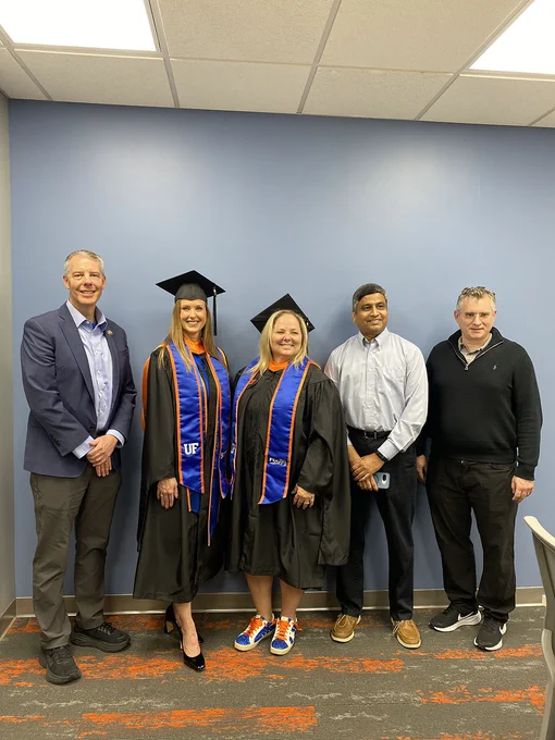 A group of five people, two wearing graduation caps and gowns with "UF" sashes, pose together indoors. Trey Tillander of FDOT is standing to the left and Amanda Carpenter and Stacy Hill are in the middle wearing graduation caps and gowns. To Stacy's right is Dr. Siva Srinivasan and next to Siva is Dr. Kyle Riding who are dressed in formal and casual attire. The setting appears to be a room with a blue wall.