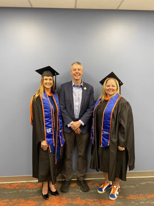 Trey Tillander of FDOT stands between Stacy Hill and Amanda Carpenter who are wearing graduation caps and gowns, featuring "UF" sashes, in front of a plain blue wall.