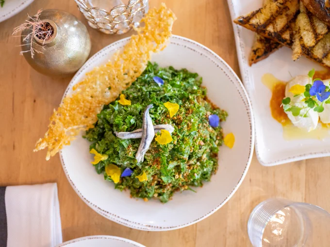 A white bowl of finely chopped kale salad topped with anchovies, yellow and blue edible flower petals, and a crispy cheese tuile, on a wooden table with grilled bread and another dish nearby.