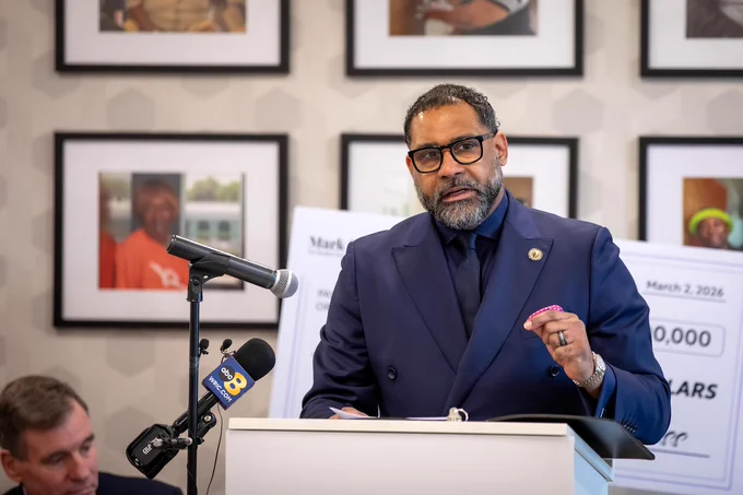Board Chair Roscoe D. Cooper III speaking at a podium with microphones during a press conference indoors.