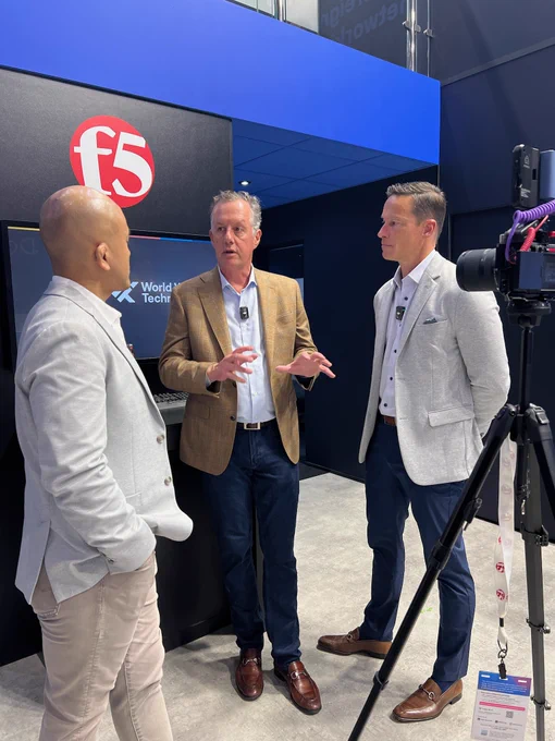 Three men in blazers stand in front of an f5 logo, engaged in conversation. One man gestures with his hands, another has his hands behind his back. A camera on a tripod is visible to the right.