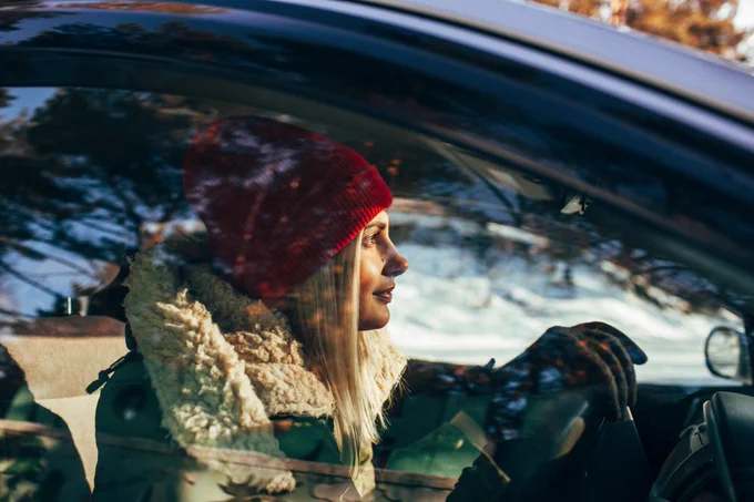 A person wearing a red beanie and a warm scarf is driving a car, with sunlight streaming through the window, creating a bright and cheerful atmosphere.