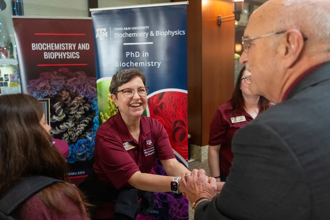 A person in a maroon Texas A&M University shirt is smiling and shaking hands with another individual. Behind them are banners for Texas A&M University, highlighting the Biochemistry and Biophysics department and a PhD program.
