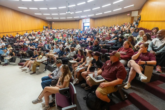 A large group of people sit in a university lecture hall with rows of tiered seating. Most seats are occupied, and attendees appear attentive, some taking notes on laptops. The lecture hall features wooden paneling and bright overhead lighting.
