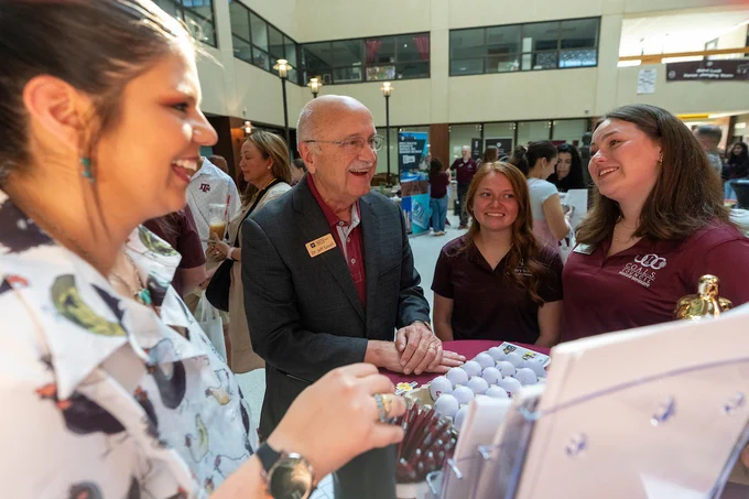 A group of individuals smiling and conversing at a professional gathering indoors, with informational materials displayed on a table. The setting appears to be an event or conference inside a building.