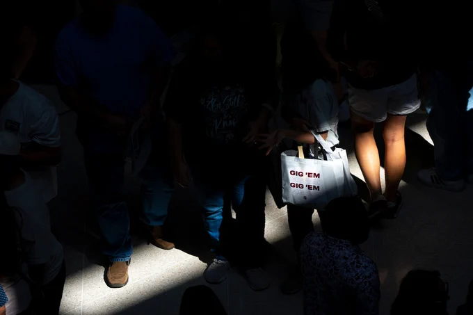 A group of people standing in a dimly lit area, with a spotlight on a tote bag. The bag has "GIG 'EM" printed in bright red letters.