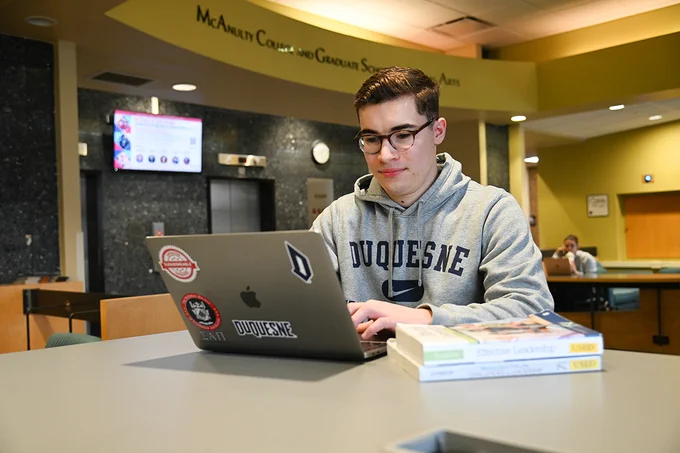 Ryan Stoudt works on his laptop while seated at a table in the foyer of College Hall. He is wearing a gray Duquesne sweatshirt and his laptop is decorated with several Duquesne-related stickers. 
