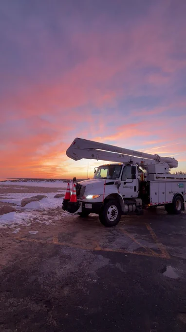 A bucket truck is parked at a snowy beach at sunset, silhouetted against a sky filled with pink, orange, and purple clouds.