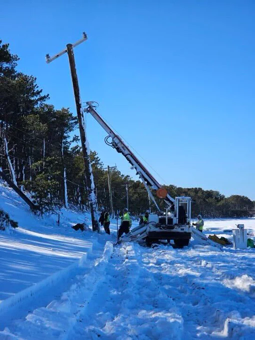 A line crew works in deep snow beside a forested roadside as a bucket truck raises a new utility pole into place. Snow blankets the ground and tree branches under a bright blue sky.