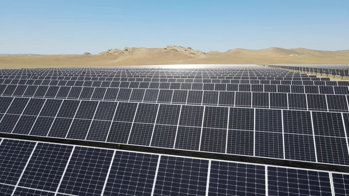Aerial view of a large utility-scale solar farm with rows of panels stretching across a desert landscape under a clear blue sky.