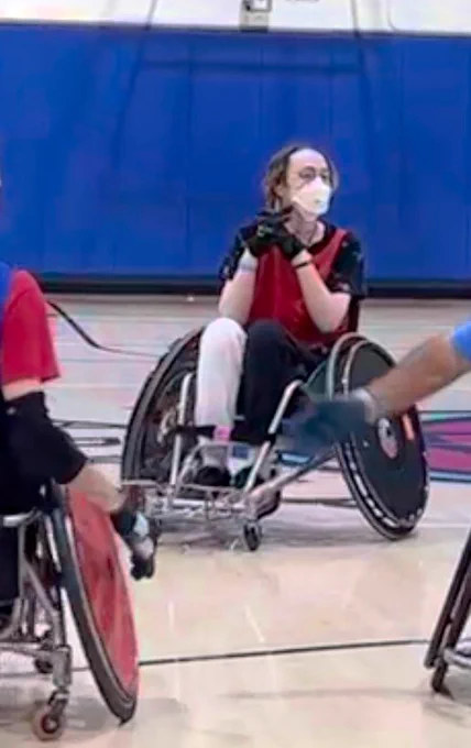Wheelchair user in a white mask and red jersey seated in a rugby wheelchair on a court with his hands clasped 