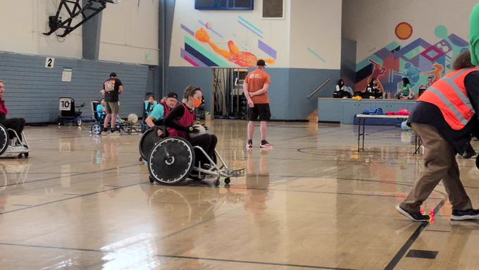 Wheelchair user in an orange mask and red jersey holding a ball whilst playing wheelchair rugby