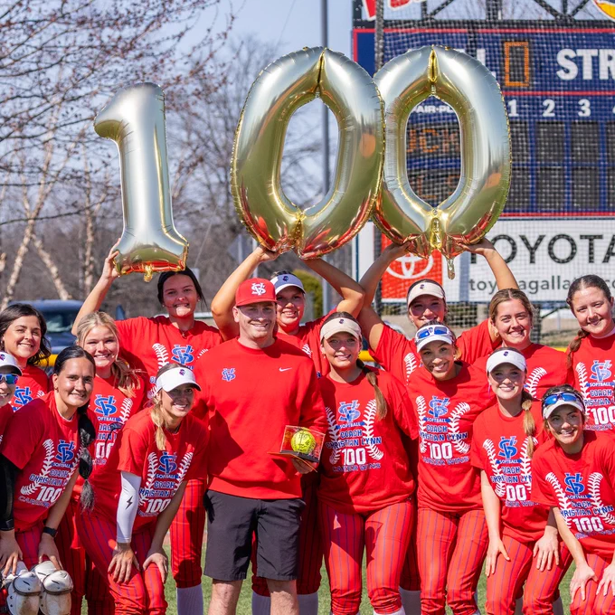 The ladies team celebrating the win for coach Lynn, holding balloons reading 100 over his head.