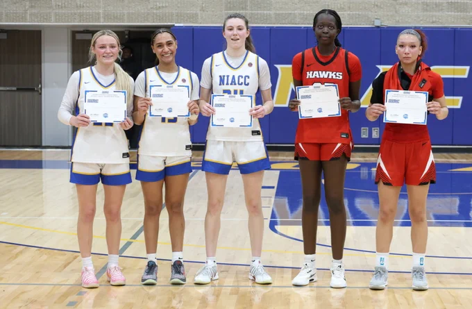 Five women’s basketball players stand on a gym court holding ICCAC All-Tournament Team certificates. Three players in white NIACC uniforms with blue and gold trim stand alongside two players in red Hawks uniforms. They pose shoulder to shoulder at center court with a purple and gold gym wall in the background.