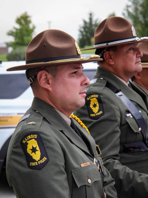 Two Illinois State Police officers in uniform stand in profile. They wear brown hats and green uniforms with "Illinois State Police" patches. A police vehicle is visible in the background.