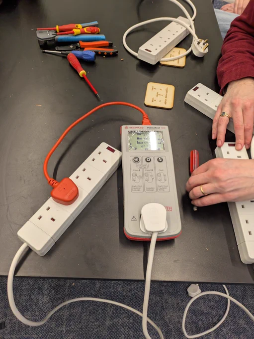 PAT tester on a table checking a newly built extension lead, with hand tools and plug-wiring kits nearby.