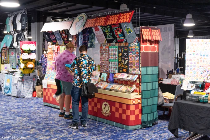 A photograph of the 2025 Dealer's Den. Two individuals are browsing a booth in the middle of the photo that is full of various furry merch.