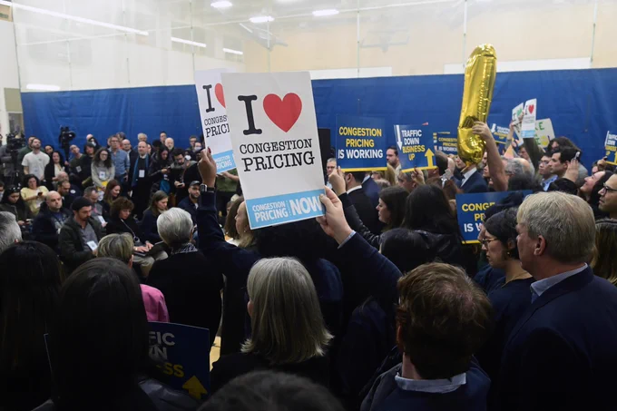 Crowd gathered indoors holding signs in support of congestion pricing. One sign prominently displays "I ♥ Congestion Pricing." Balloons and a microphone setup are visible.