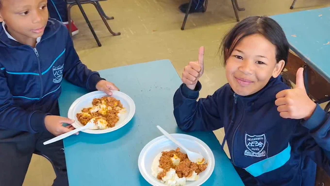 Two South African school children in blue school uniforms sit at a table enjoying the nutritious meal. One gives the thumbs up sign.